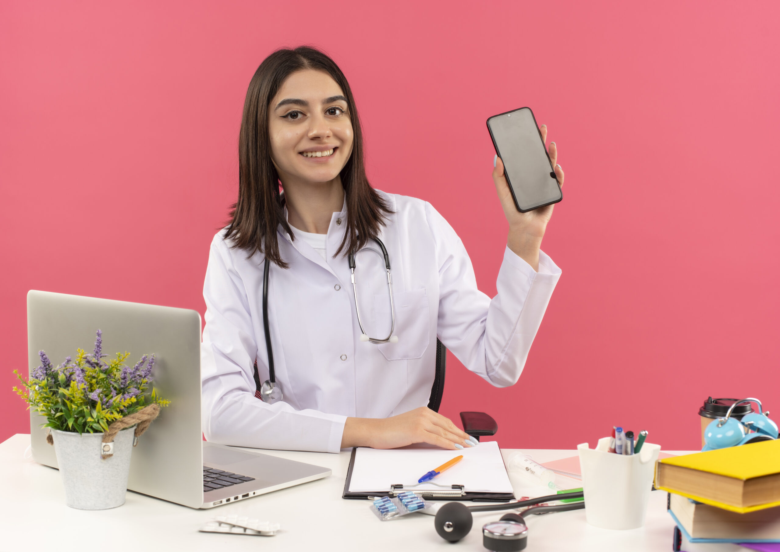 young female doctor white coat with stethoscope around her neck showing smartphone smiling confident sitting table with laptop pink wall scaled